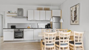 A modern kitchen with white cabinets, a built-in oven, and a dining area with wooden chairs and a table. - Gudhjem Søpark Lejl. 16 (Gudhjem)