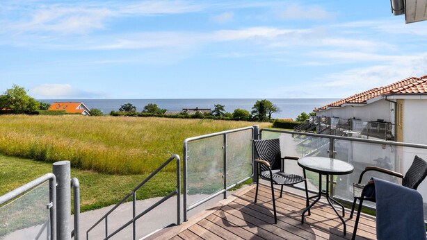 A balcony with a view of the sea, a grassy field, and a building with a tiled roof. - Gudhjem Søpark Lejl. 16 (Gudhjem)