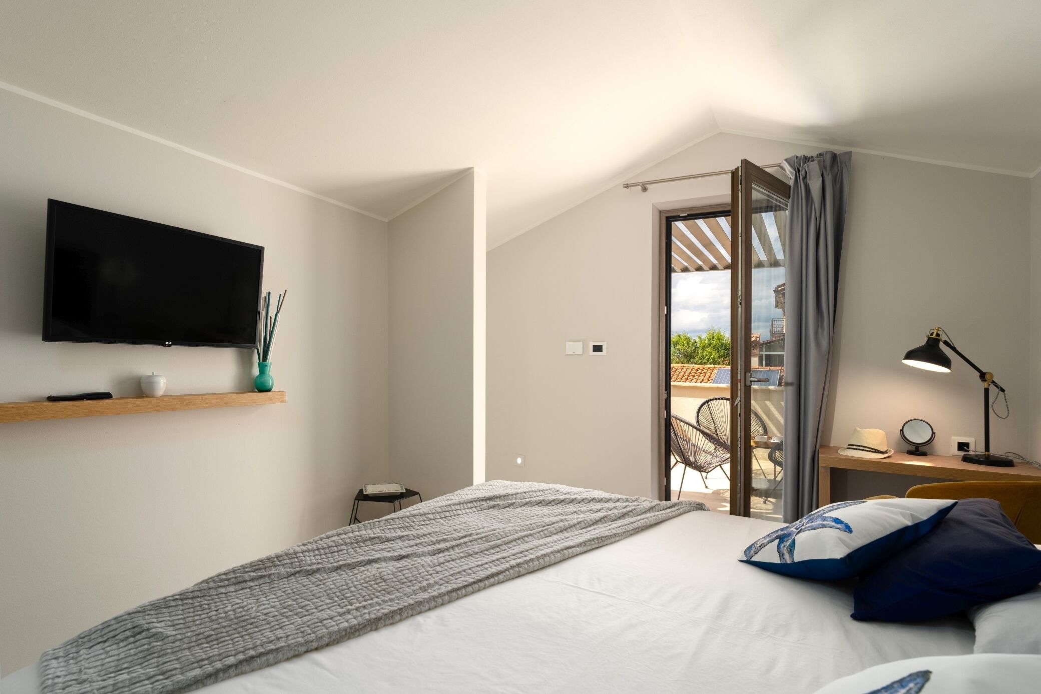 Habitació d'hotel moderna amb un llit gran, televisió de pantalla plana penjada a la paret, una taula petita amb una làmpada, i vista al pati exterior per una porta oberta.