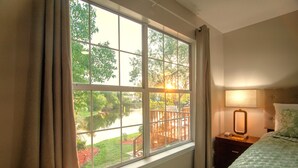 Ein Hotelzimmer mit großem Fenster, einem Nachttisch mit Lampe und einem Bett mit grüner Steudecke.