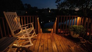 Eine Holzterrasse mit Schaukelstuhl und Topfpflanze, die zu einem Gewässer im Abendlicht führt.