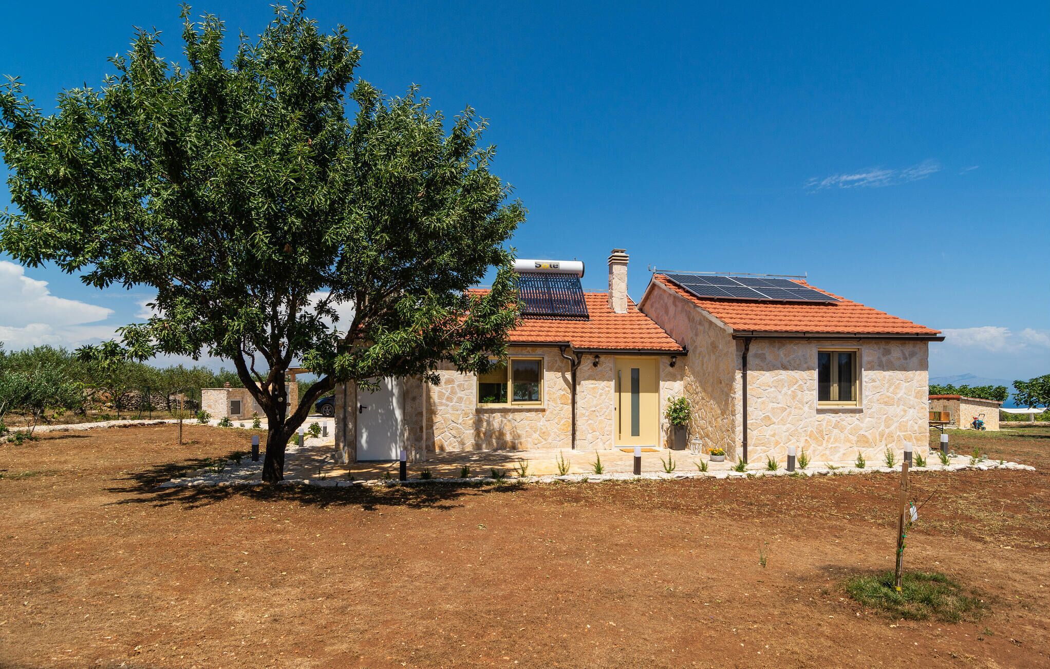 A stone house with a red-tiled roof, solar panels, and a large tree in front.