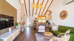 Ein modernes Wohnzimmer mit einem grünen Sofa, Korbstühlen, einem Fernseher und einer Küchenzeile mit Holzbalken und einem Fenster.