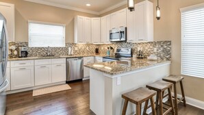 A kitchen with white cabinets, a granite countertop, a stainless steel dishwasher, and a microwave. There are wooden stools and a window with blinds. - Flamingo Beach Hideaway (St. Augustine)