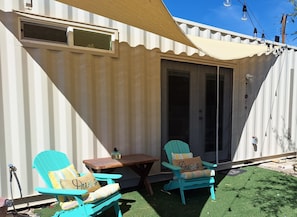 A covered outdoor seating area with two chairs and a table, a door, and a window.