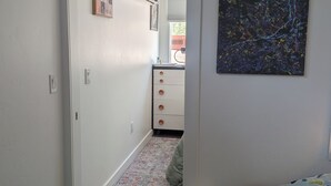 A narrow hallway with a patterned rug, a white wall with a mounted air conditioner, a framed collage, and a small round stool.