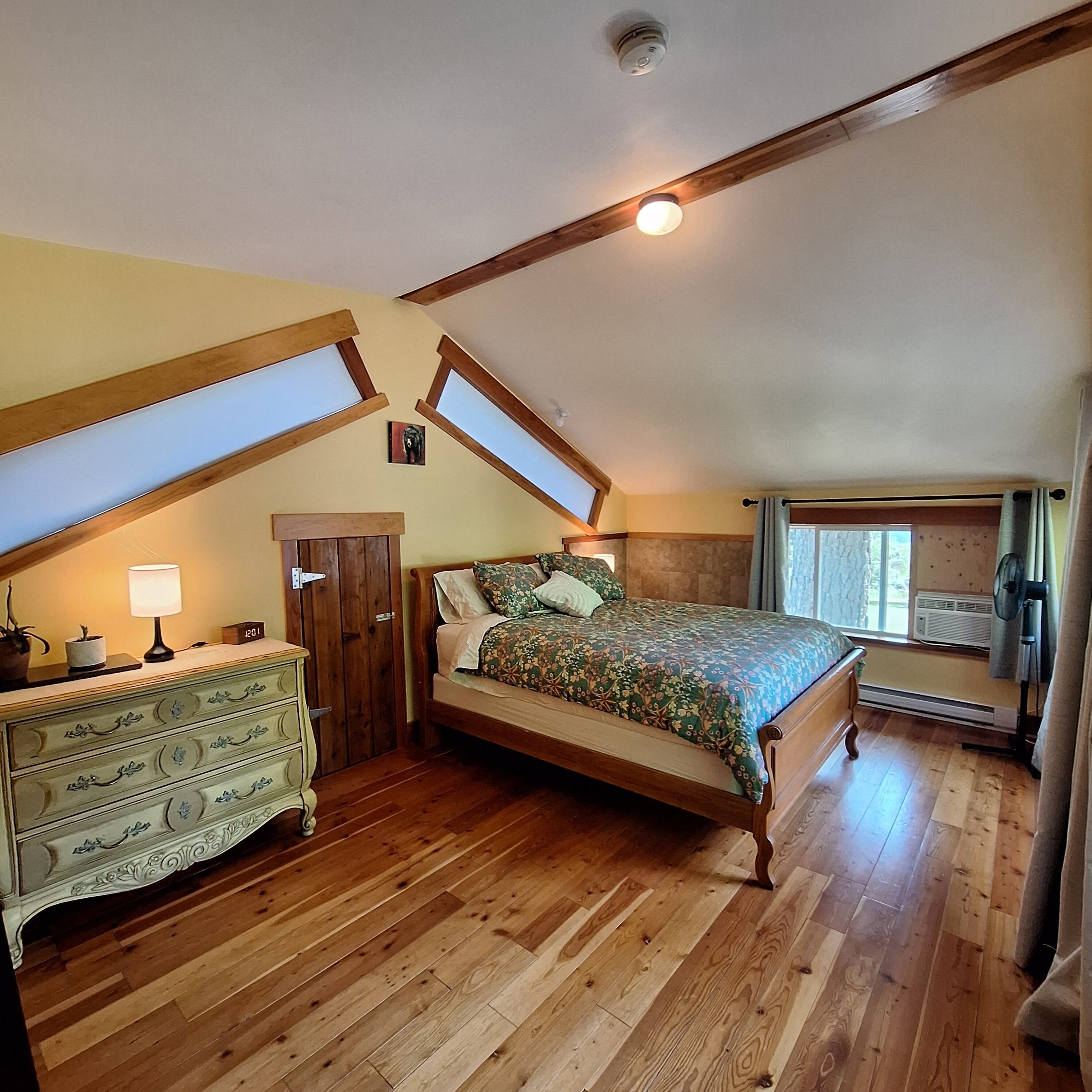 Une chambre avec un plafond en pente, des poutres en bois, un lit avec une literie à motifs floraux, une commode, une lampe et une fenêtre avec des rideaux.