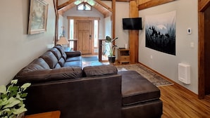 Una sala de estar acogedora con un sofá de cuero marrón, vigas de techo de madera, un televisor montado y un cuadro grande de un bosque.