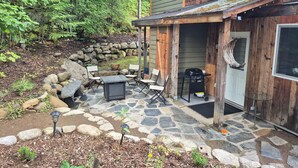 Una casa de madera con un patio de piedra, mobiliario exterior y una parrilla.