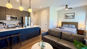 Quarto de hotel moderno com cozinha compacta, sofá, cama, ventoinha no teto e uma moldura na parede.