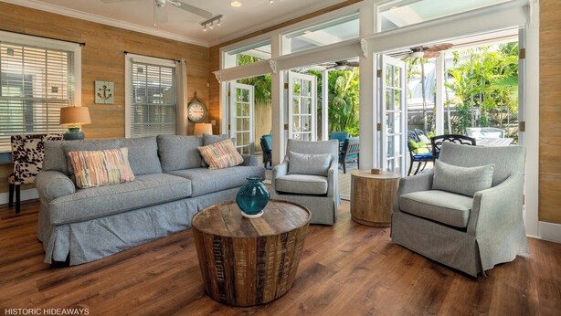 A spacious living room with a grey sofa, two armchairs, a coffee table, and a ceiling fan. Large windows provide ample natural light and a view of greenery outside. - Boomerang Boathouse (Key West)