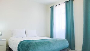 Een slaapkamer met een bed, witte kussens, een blauwgroene deken, een nachtkastje en blauwgroene gordijnen.
