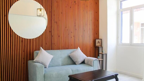 Een kamer met een lichtblauwe bank, een donkerhouten salontafel, een ronde spiegel en een wand van houten latten.