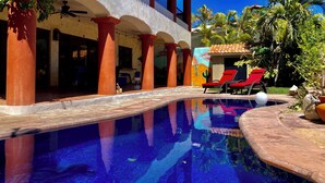 A pool area with red lounge chairs, a clear blue sky, and a building with red pillars and a mural. - Charming 5-bed house with sweeping ocean views in delightful San Jose Del Cabo. (San Jose Del Cabo)