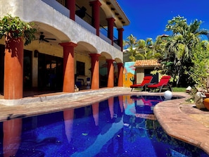A pool area with red lounge chairs, a clear blue sky, and a building with red pillars and a mural. - Charming 5-bed house with sweeping ocean views in delightful San Jose Del Cabo. (San Jose Del Cabo)