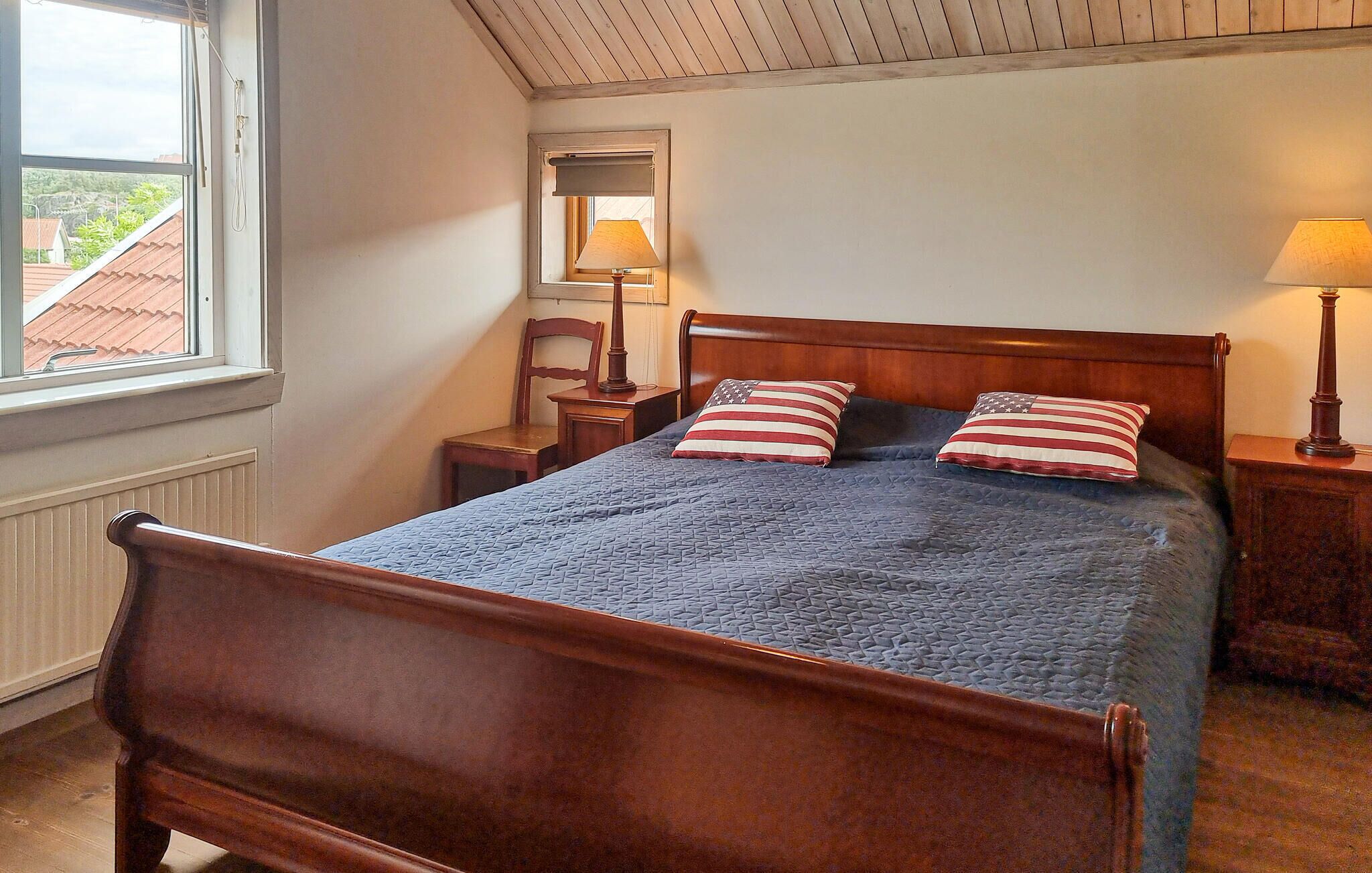Et soveværelse med en træseng, to puder med et amerikansk flag-design, et natbord med en lampe, en stol og et vindue med udsigt til bygninger.