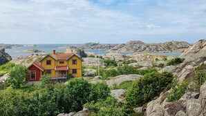 A yellow house with a red door and a balcony, surrounded by rocky terrain and greenery, overlooking a coastal area. - Cozy home in Tjörn with sauna (Tjörn)