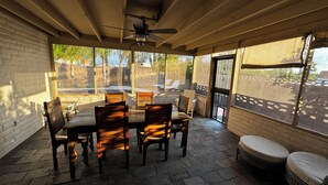 En overdækket terrasse med et bord og stole, en ventilator i loftet og udsigt til baghaven.