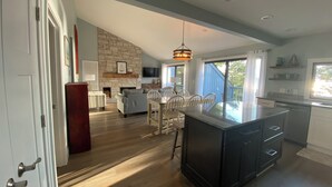 A spacious kitchen with a large island, a dining area with white chairs, a fireplace, and a stone accent wall. - Sleeping Bear Beauty! Fully Remodeled Beachfront Condo with summer availability  (Glen Arbor)
