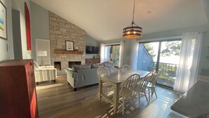 A spacious living area with a stone fireplace, a dining table with chairs, a sofa, and a television mounted on the wall. - Sleeping Bear Beauty! Fully Remodeled Beachfront Condo with summer availability  (Glen Arbor)