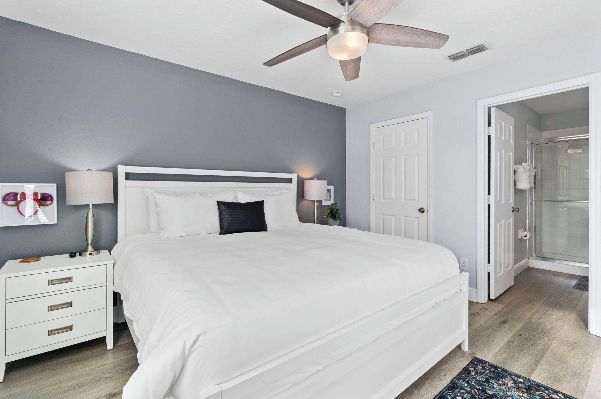 A bedroom with a white bed, a nightstand, a lamp, a ceiling fan, and a door leading to a bathroom.