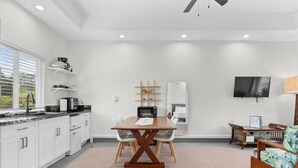 A modern kitchen with white cabinets, a wooden dining table, and a TV mounted on the wall. - Red Ti Cottage   (Keaau)