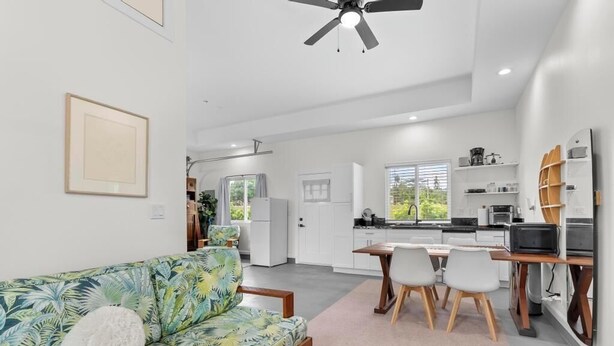 A modern kitchen with white cabinets, a dining area with a table and chairs, and a living room with a sofa and a ceiling fan. - Red Ti Cottage   (Keaau)