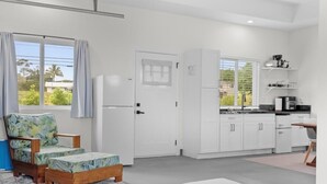 A modern kitchen with white cabinets, a dining area with a wooden table, and a view of greenery outside. - Red Ti Cottage   (Keaau)