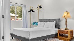 A neatly arranged bedroom with a bed, a bedside table with a lamp, a shoe rack, and a view through a window. - Red Ti Cottage   (Keaau)