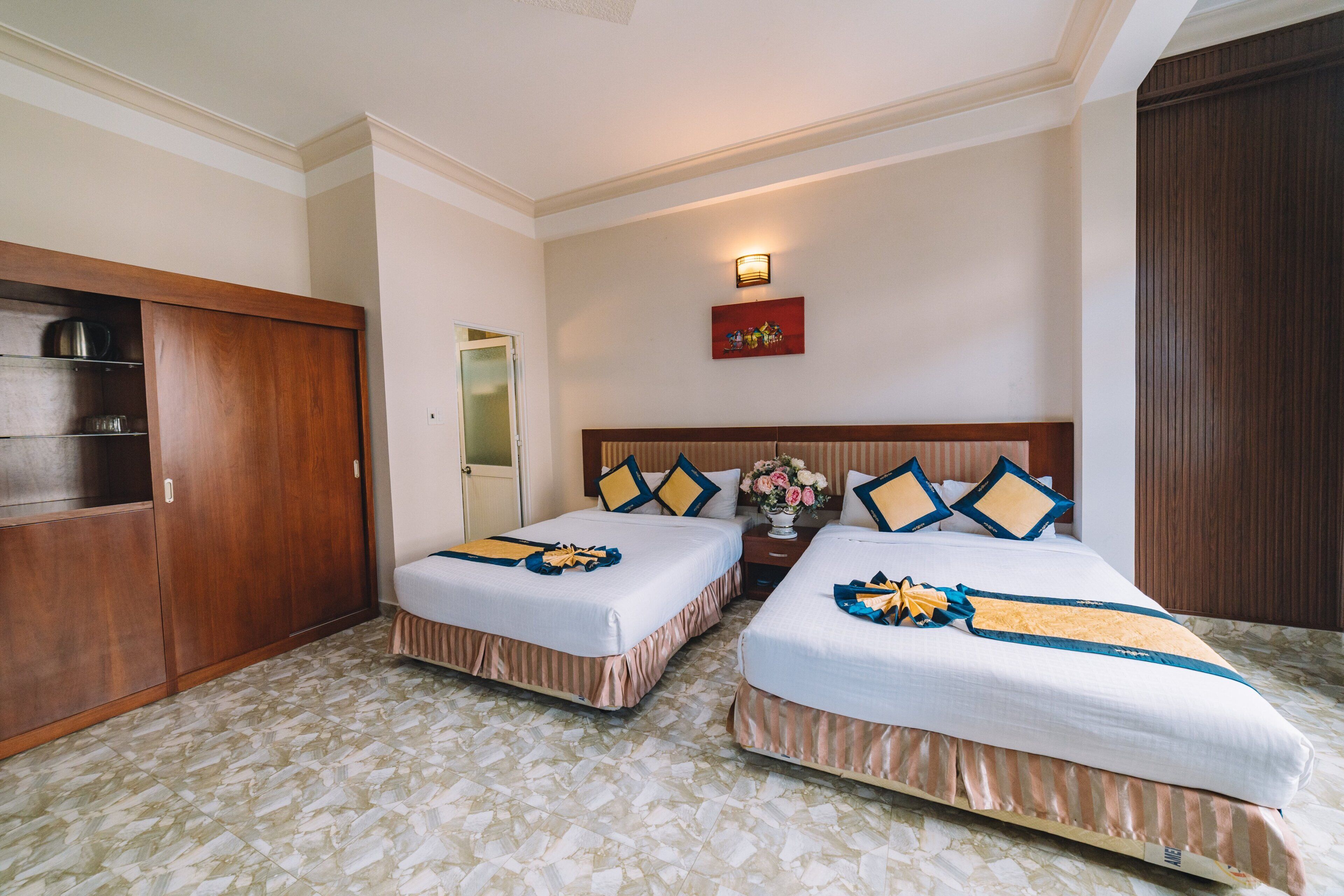 A hotel room with two beds, a wardrobe, a nightstand with a vase of flowers, and a wall-mounted light fixture.