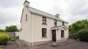 A two-story house with a gabled roof, a front porch, and a small garden area. - Countryside Retreat Dingle (Municipal District of Castleisland-Corca Dhuibhne)