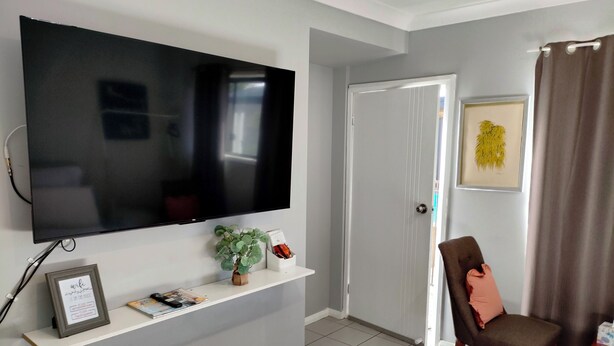 Een woonkamer met een flatscreen tv aan de muur, een klein plantje op een plank, een lijst met een foto en een fauteuil met een kussen.