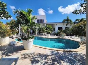 A pool area with a modern house in the background, surrounded by potted plants and a clear blue sky. - First Day Hotel on Aruba (Oranjestad)
