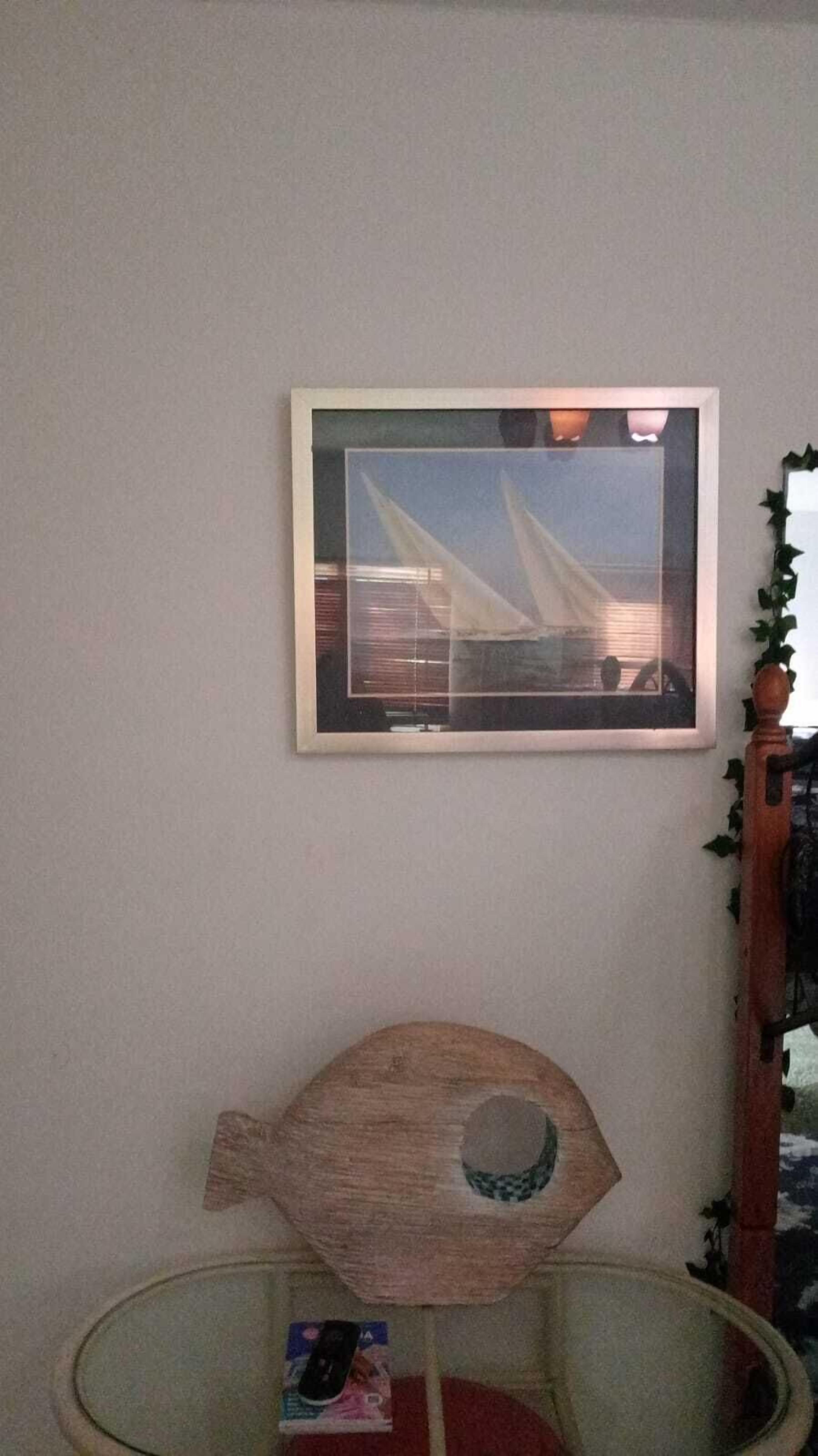 Un cuadro con un velero en la pared, un adorno de madera con forma de pez y una mesa de cristal con una revista.
