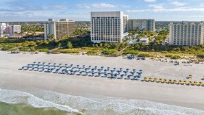 Ein Strand mit Reihen aus blauen und gelben Sonnenschirmen, ein großes Gebäude mit einer rasterartigen Fassade und weitere Hochhäuser im Hintergrund.