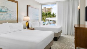 Ein Hotelzimmer mit zwei Betten, einem Schreibtisch, einer Lampe und einem Fernseher. Es gibt ein Fenster mit Blick auf einen Pool und Palmen.