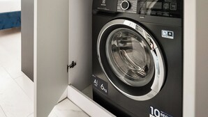 A compact, modern laundry room with a front-loading washing machine, a shelf with folded towels, and a yellow basin. - Plac Politechniki Cosy Studio (Warszawa)