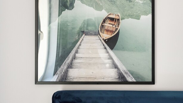 A framed artwork of a wooden dock leading to a boat on calm water with mountains in the background. - Plac Politechniki Cosy Studio (Warszawa)