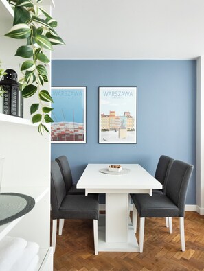 A dining area with a white table and grey upholstered chairs, two framed cityscape posters on the wall, and a potted plant on the left.