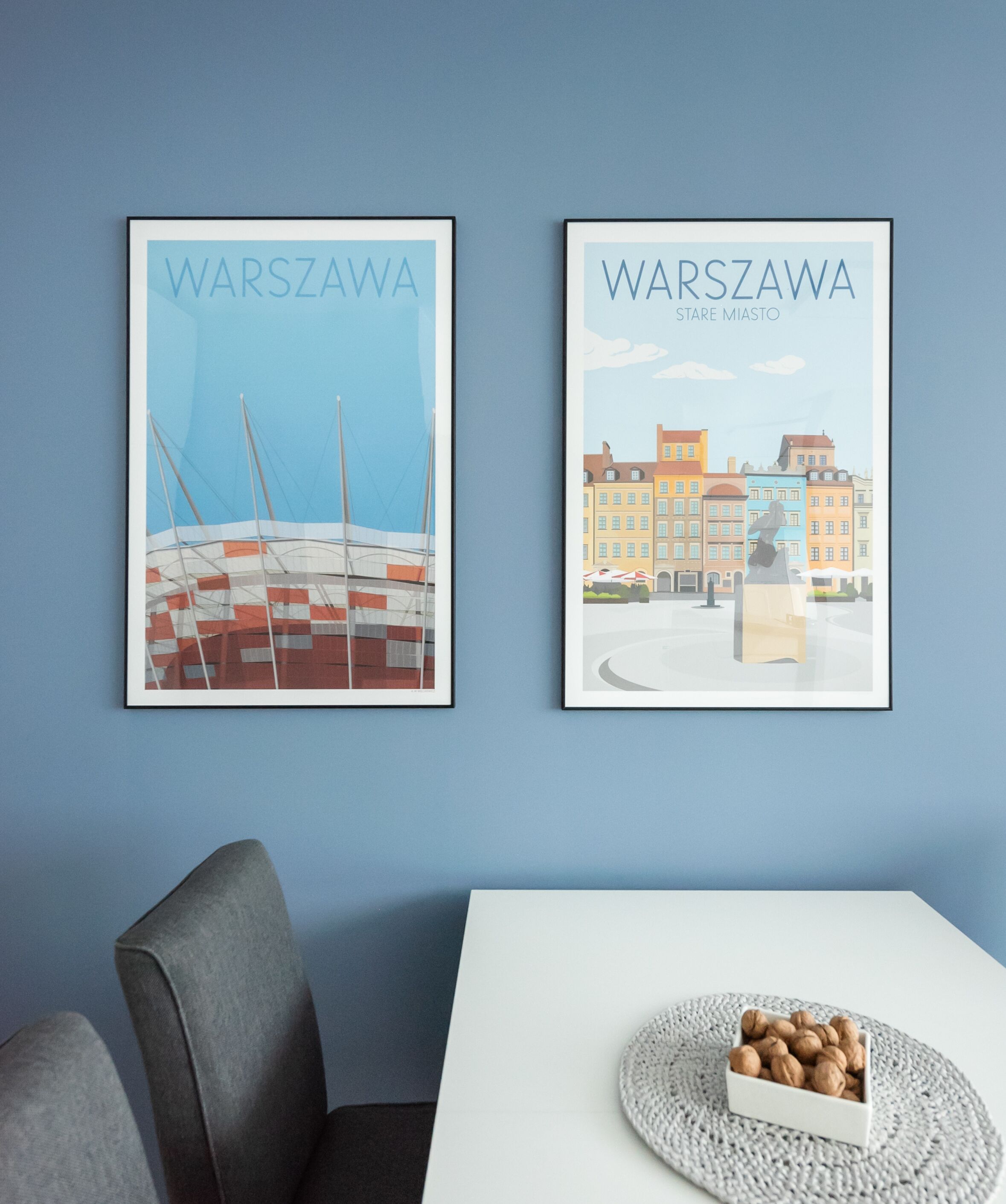 Dois cartazes emoldurados de Varsóvia numa parede azul, uma cadeira cinzenta e uma mesa com um apoio de mesa trançado e uma tigela de frutos secos.