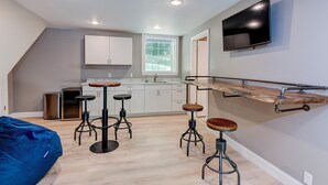A modern kitchen with white cabinets, a large window, and a built-in TV. There are four stools and a blue bean bag chair.
