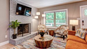 A living room with a brick fireplace, a flat-screen TV mounted above it, two patterned armchairs, a round coffee table, and a large window with a view of trees.