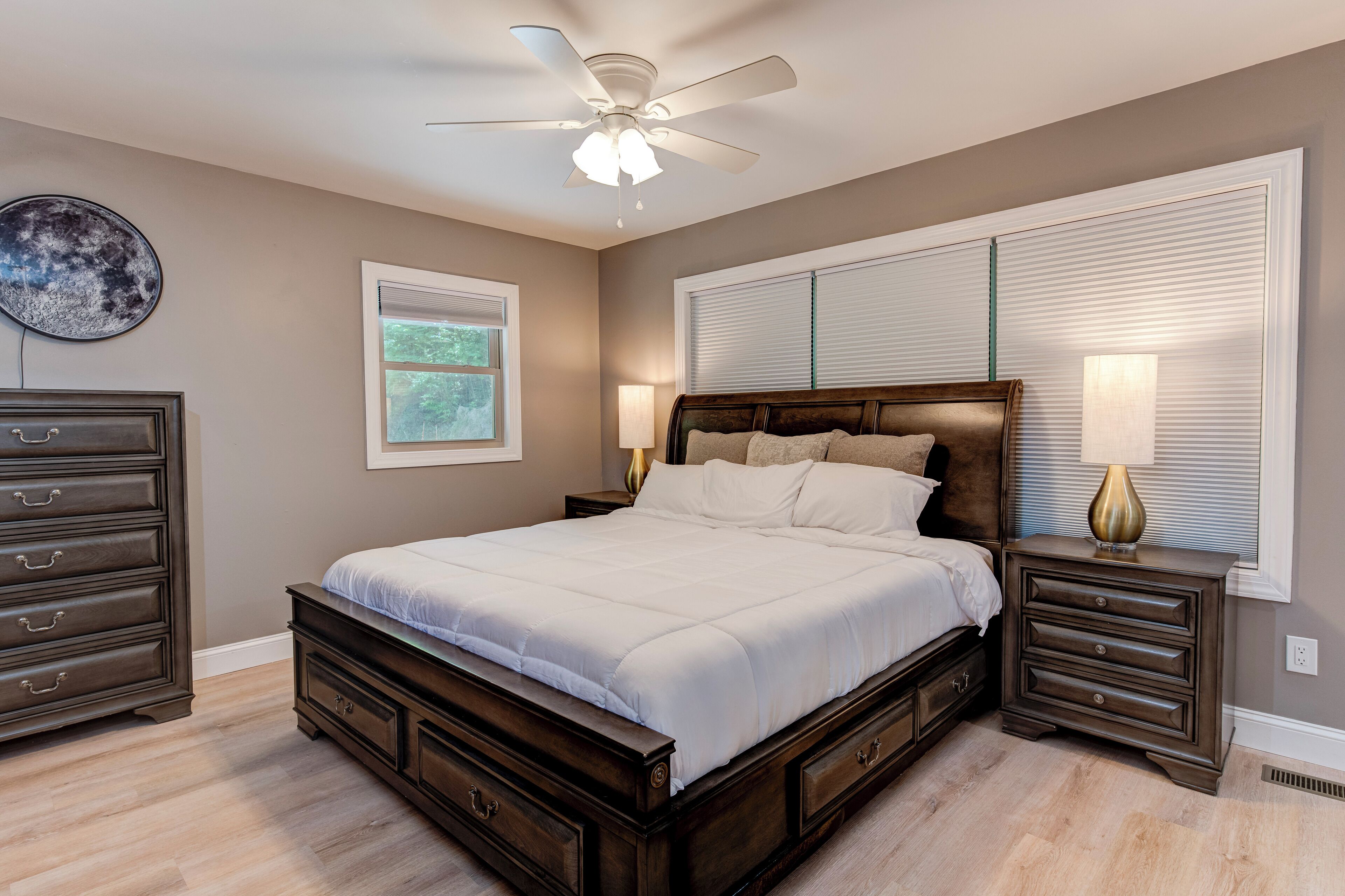 A bedroom with a large bed, a ceiling fan, a dresser, a nightstand, and a window with blinds.