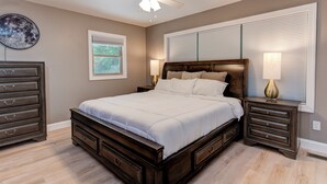 A bedroom with a large bed, a ceiling fan, a dresser, a nightstand, and a window with blinds.