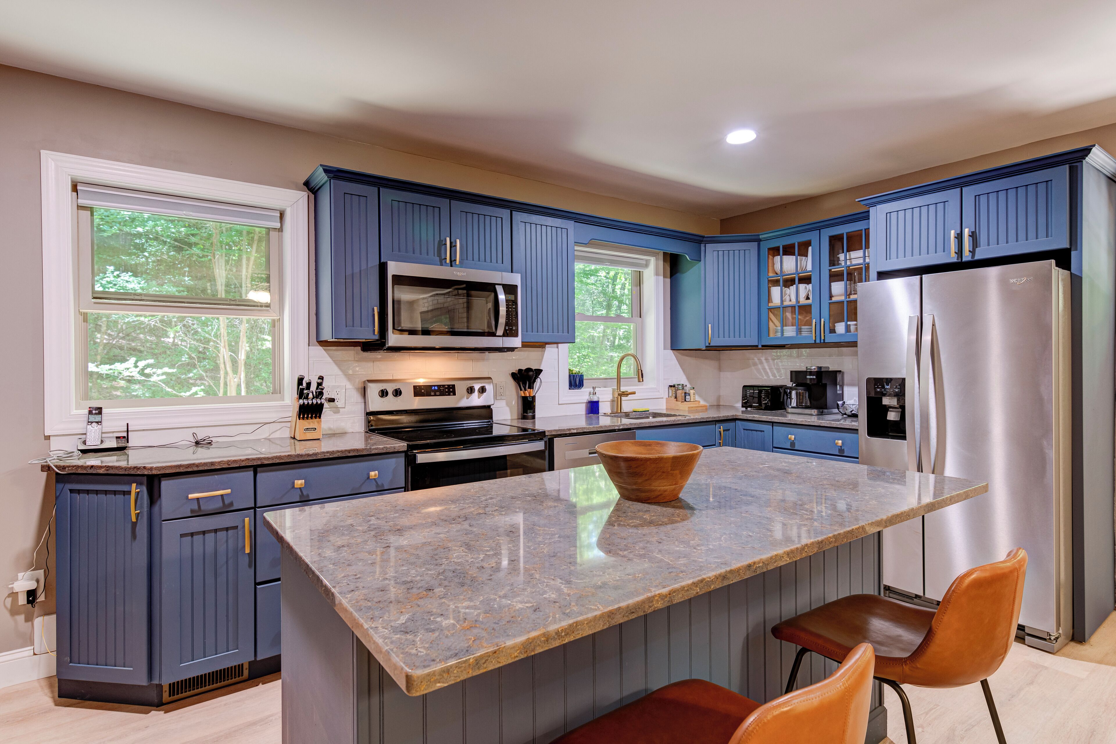 A modern kitchen with blue cabinets, stainless steel appliances, a granite island, and a view of trees through the windows.