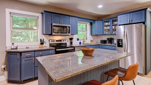 A modern kitchen with blue cabinets, stainless steel appliances, a granite island, and a view of trees through the windows.