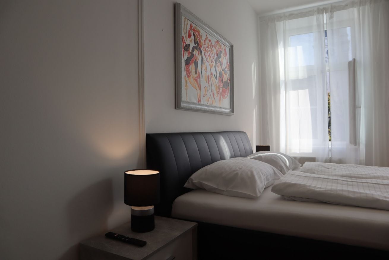 A hotel room with a bed, a nightstand with a lamp, a remote control, and a framed artwork on the wall.
