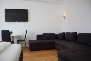 A modern living room with a sectional sofa, a flat-screen TV mounted on the wall, a small table with a chair, and a potted plant.