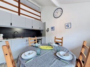 Ein Essbereich mit einem runden Tisch, Stühlen, Tellern, Gläsern und Besteck. An der Wand hängt eine Uhr und im Hintergrund ist eine Küche zu sehen.