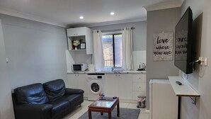 Een gezellige, compacte leefruimte met een bank, een salontafel, een wasmachine, een kleine koelkast en een aan de muur bevestigde televisie.